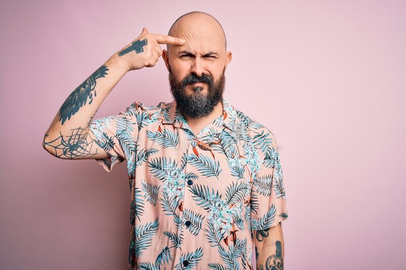 Handsome bald man beard tattoo wearing casual floral shirt over pink background pointing unhap
