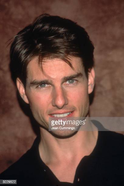 Headshot of american actor tom cruise smiling in a black shirt in front of a brown backdrop