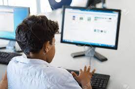 Rear View Of Multiethnic Boy Using Desktop Computer In School Library. Back  View Of Mixed Race Child Working In Classroom With Computer At Elementary  School. Young Arab Schoolboy Typing On Keyboard. Stock