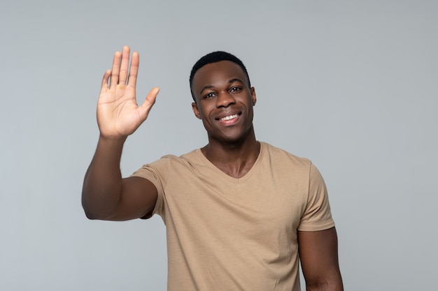 Greeting happy smiling young dark skinned man raising hand greeting standing isolated light gr