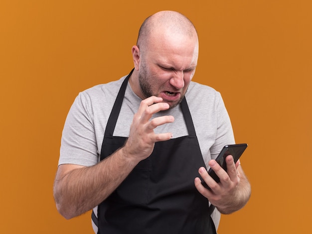Angry slavic middle aged male barber uniform holding looking phone isolated orange wall 141793