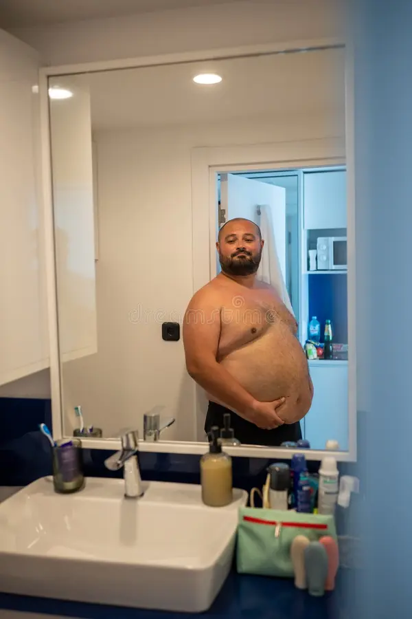 Overweight smiling man naked torso suffering extra weight looks mirror touches abdomen lookin